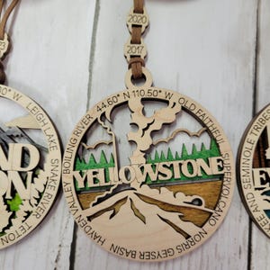 Puede incluir: Adorno de madera con un mapa detallado del Parque Nacional de Yellowstone con las palabras "Yellowstone" y las coordenadas "44.60&deg; N 110.50&deg; W" inscritas. El adorno est&aacute; decorado con pintura verde, marr&oacute;n y blanca.