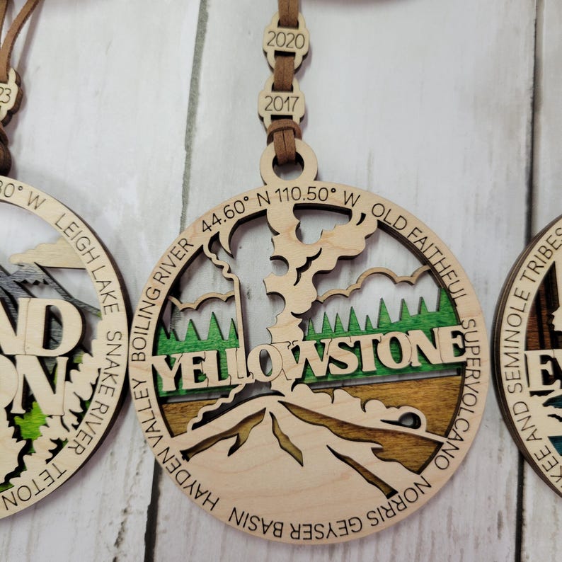 Puede incluir: Adorno de madera con un mapa detallado del Parque Nacional de Yellowstone con las palabras "Yellowstone" y las coordenadas "44.60&deg; N 110.50&deg; W" inscritas. El adorno est&aacute; decorado con pintura verde, marr&oacute;n y blanca.