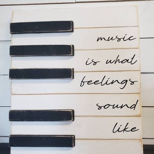 Puede incluir: Un letrero de madera dise&ntilde;ado para parecerse a las teclas de un piano. El letrero es blanco con acentos rectangulares negros. Las palabras "music is what feelings sound like" est&aacute;n escritas en escritura negra.