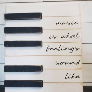 Puede incluir: Un letrero de madera blanco con teclas de piano negras y las palabras "music is what feelings sound like" en escritura negra. El letrero es rectangular y tiene un aspecto r&uacute;stico y envejecido.