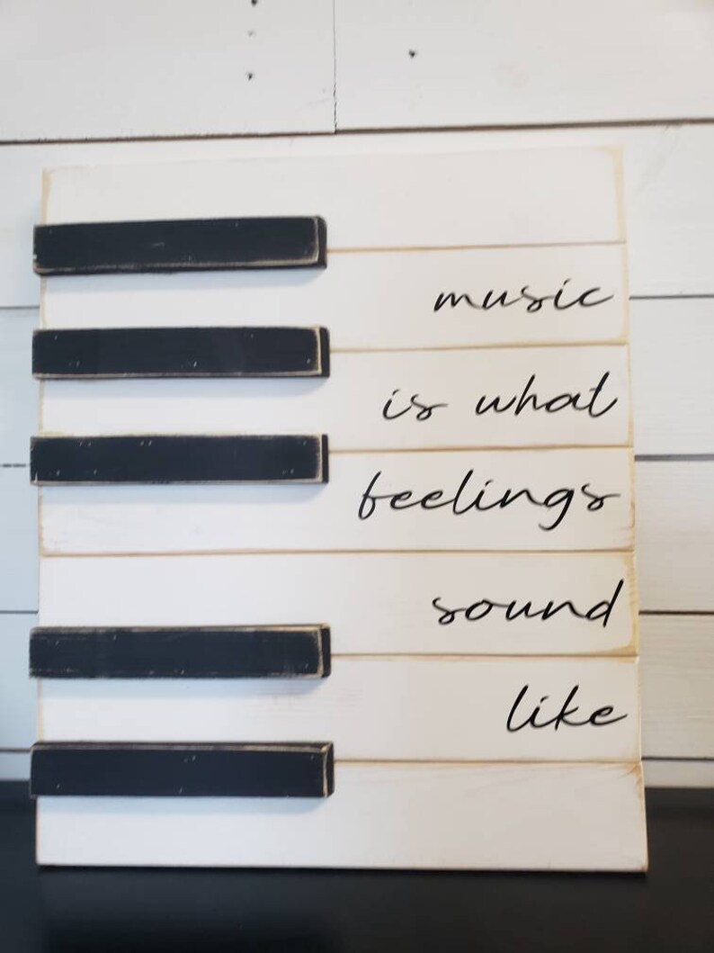 Puede incluir: Un letrero de madera blanco con teclas de piano negras y las palabras "music is what feelings sound like" en escritura negra. El letrero es rectangular y tiene un aspecto r&uacute;stico y envejecido.