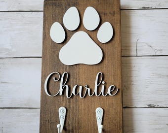 Puede incluir: Un soporte de correa para perro de madera marrón con una huella de pata blanca y el nombre "Charlie" en letras blancas. Dos ganchos blancos están unidos a la parte inferior del soporte.