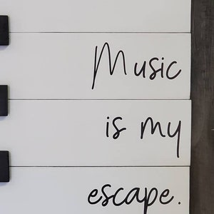 Puede incluir: Letrero de madera blanco con las palabras "Music is my escape" en letras cursivas negras. El letrero est&aacute; hecho de tablones de madera horizontales con acentos cuadrados negros. Una pieza decorativa para un amante de la m&uacute;sica.