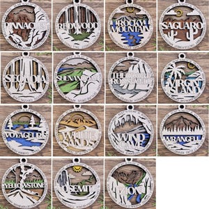 Puede incluir: Doce adornos redondos de madera, cada uno con un parque nacional diferente de los Estados Unidos. Los adornos est&aacute;n pintados con varios colores y detalles, incluyendo monta&ntilde;as, &aacute;rboles, agua y nombres de parques.