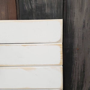 Puede incluir: Primer plano de un letrero de madera blanco con un aspecto envejecido. El letrero est&aacute; hecho de tablones horizontales con una apariencia ligeramente desgastada. Los bordes tienen un color marr&oacute;n claro y el fondo es de madera oscura.