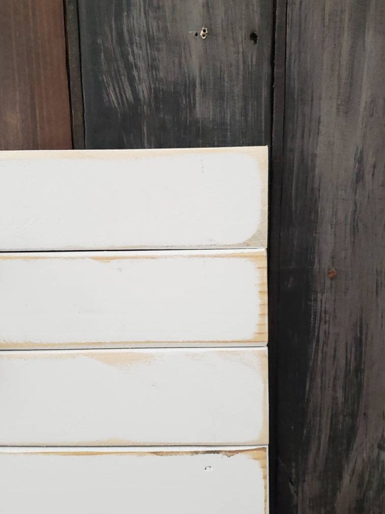 Puede incluir: Primer plano de un letrero de madera blanco con un aspecto envejecido. El letrero est&aacute; hecho de tablones horizontales con una apariencia ligeramente desgastada. Los bordes tienen un color marr&oacute;n claro y el fondo es de madera oscura.