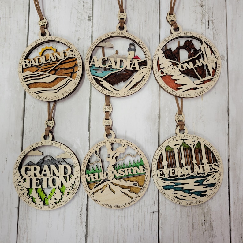 Puede incluir: Seis adornos de madera, cada uno con un parque nacional diferente. Los adornos son redondos y tienen un cord&oacute;n marr&oacute;n para colgar. Los parques que se muestran son Badlands, Acadia, Canyonlands, Grand Teton, Yellowstone y Everglades. Cada adorno incluye el nombre del parque, las coordenadas de la ubicaci&oacute;n y una peque&ntilde;a ilustraci&oacute;n de un hito o caracter&iacute;stica del parque.