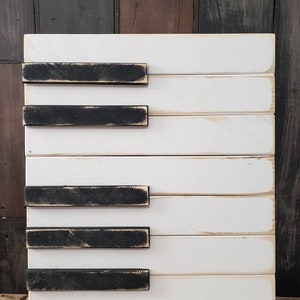 Puede incluir: Arte de pared con teclas de piano de madera. La pieza presenta tablas de madera blancas con teclas negras desgastadas. La obra rectangular tiene un estilo r&uacute;stico. El fondo es una mezcla de tablas de madera, creando un ambiente de granja.