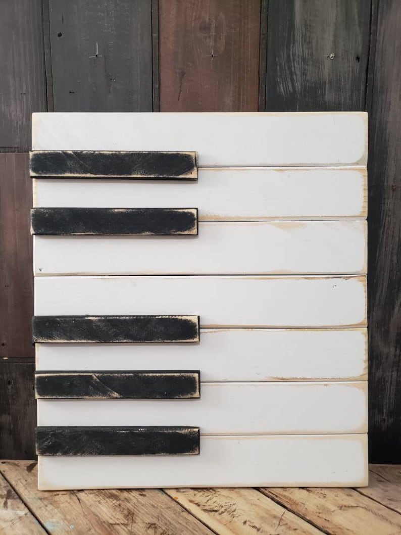 Puede incluir: Arte de pared con teclas de piano de madera. La pieza presenta tablas de madera blancas con teclas negras desgastadas. La obra rectangular tiene un estilo r&uacute;stico. El fondo es una mezcla de tablas de madera, creando un ambiente de granja.