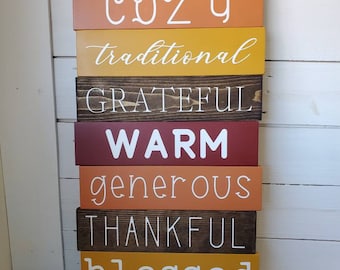 Puede incluir: Una pila de letreros de madera con varias palabras en letras blancas. Los letreros son en tonos de marrón, naranja, amarillo y rojo. El letrero superior dice "Welcome to our", seguido de "COZY", "traditional", "GRATEFUL", "WARM", "generous", "THANKFUL", "blessed" y "HOME".