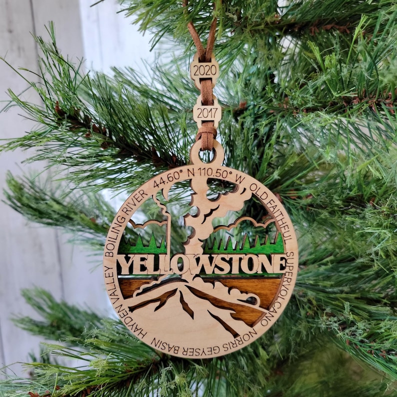 Puede incluir: Adorno de Navidad de madera con una ilustraci&oacute;n detallada del Parque Nacional de Yellowstone, incluyendo el g&eacute;iser Old Faithful, el valle de Hayden y la cuenca de g&eacute;iseres de Norris. El adorno es marr&oacute;n con detalles verdes e incluye el texto "Yellowstone" y las coordenadas "44.60&deg; N 110.50&deg; W".