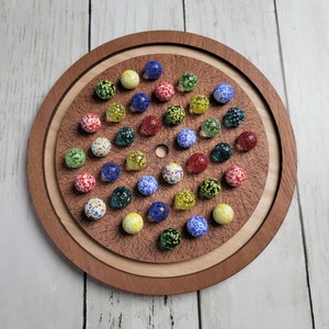 May include: A wooden game board with a circular mandala design and a ring of colorful marbles around the center. The marbles are arranged in a circle and are of various colors, including red, blue, green, yellow, and white.
