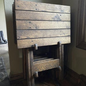 May include: A wooden folding table with a natural finish. The tabletop is made of horizontal planks, and the legs are designed to fold for storage. The table is a warm brown color and appears to be made of solid wood.