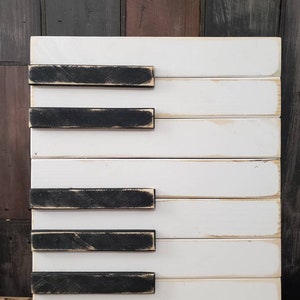 Puede incluir: Una pieza de arte mural de teclas de piano de madera. El arte presenta tablones de madera blancos con teclas rectangulares negras. La pieza tiene una apariencia envejecida y r&uacute;stica, con vetas de madera visibles y un acabado desgastado.