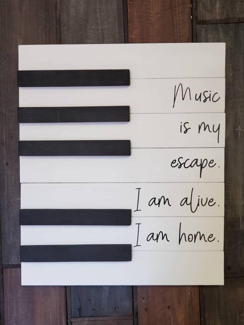 Puede incluir: Un letrero de madera blanco con detalles en negro dise&ntilde;ado para parecerse a las teclas de un piano. El letrero presenta las frases "Music is my escape. I am alive. I am home." en escritura negra, creando un tema musical.