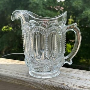 May include: Clear glass pitcher with a decorative, embossed pattern. The pitcher has a handle and a spout for easy pouring. The design includes vertical panels and floral accents. The pitcher is sitting on a wooden surface.