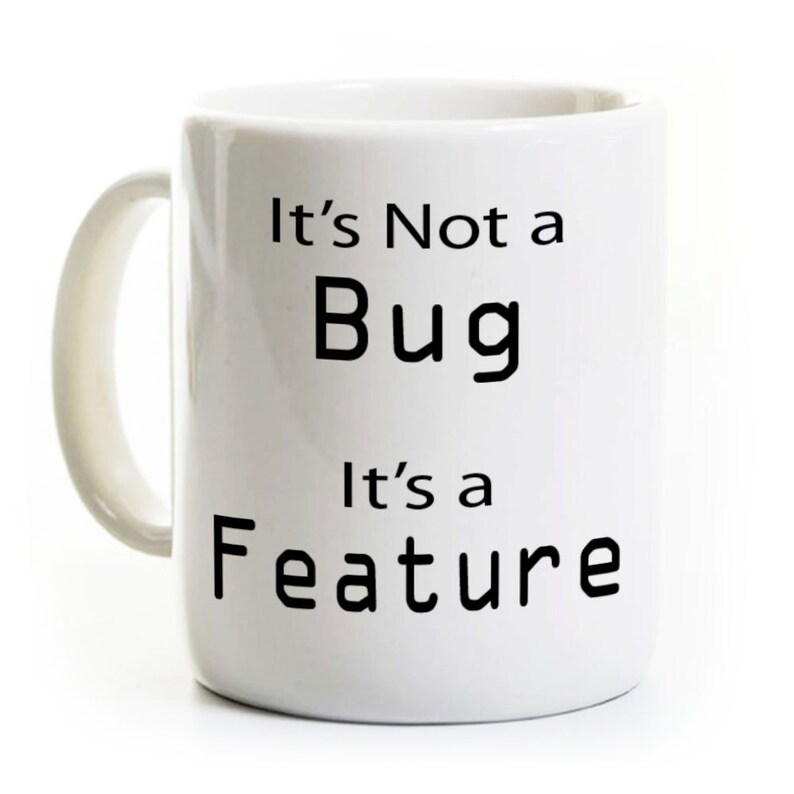 Op de afbeelding: Witte keramische mok met een gebogen handvat. De mok heeft de tekst "It's Not a Bug, It's a Feature" in zwarte, vette letters. De mok is ontworpen voor warme dranken.