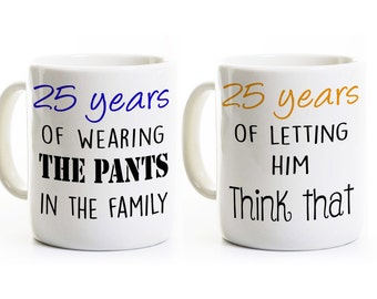 May include: Two white coffee mugs with black text. The mug on the left says "25 years of wearing the pants in the family" in blue text. The mug on the right says "25 years of letting him think that" in orange text.