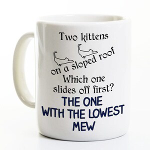 May include: White ceramic mug with a handle, featuring the text "Two kittens on a sloped roof. Which one slides off first? THE ONE WITH THE LOWEST MEW" in black and blue lettering, with two cat illustrations.