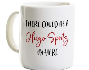 May include: A white ceramic mug with a handle on the left side. The front of the mug features black printed text that reads "THERE COULD BE A" and "IN HERE" with the red cursive script text "Hugo Spritz" positioned in the center of the lines.