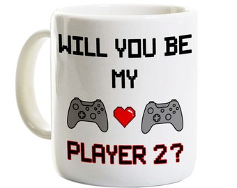 May include: White ceramic mug with the text "WILL YOU BE MY PLAYER 2?" in black and red pixelated font. The design includes two gray game controllers and a red pixel heart.