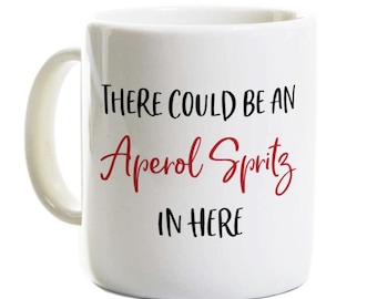 May include: A white ceramic mug with a handle featuring the black printed text "THERE COULD BE AN" and "IN HERE" with the red script text "Aperol Spritz" centered between the two lines of black text on the front of the mug.