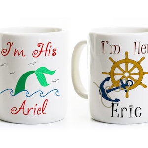 Puede incluir: Dos tazas de cer&aacute;mica blanca con asas. La taza de la izquierda tiene una cola de sirena verde con el texto "I'm His Ariel". La taza de la derecha tiene un ancla azul y un tim&oacute;n dorado con el texto "I'm Her Eric".