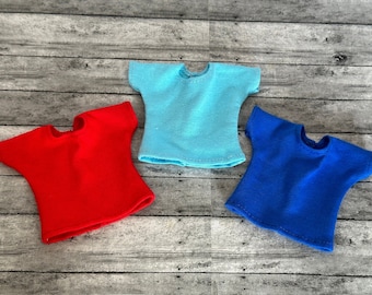 May include: Three plain t-shirts in red, blue, and light blue. The shirts are laid out on a woodgrain surface.