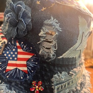 May include: Denim bucket hat with patriotic embellishments. Features a star-spangled banner star, a blue denim flower, and a beaded flower. The hat has a distressed, frayed edge and a blue star pattern.