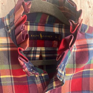 May include: A long-sleeve button-down shirt featuring a multi-colored plaid pattern in red, blue, green, yellow, and white. The shirt has a high, ruffled collar and a black interior neck label that reads Ralph Lauren with a size L.
