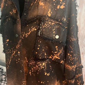 May include: Black denim jacket with orange bleach splatter effect. The jacket features a button-up front, two chest pockets, and frayed edges. The distressed design gives it a unique, vintage look. A stylish, casual outerwear piece.