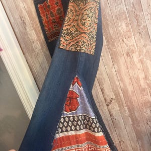 May include: Blue denim jeans with decorative patches. The patches feature intricate patterns in shades of red, orange, and olive green. The jeans have a unique, patchwork design.
