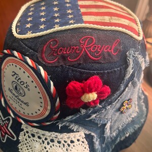 May include: Denim baseball cap with an American flag design, the Crown Royal logo, and a Tito's Vodka patch. Features a red crocheted flower and a white lace trim. The cap is a patchwork of denim.