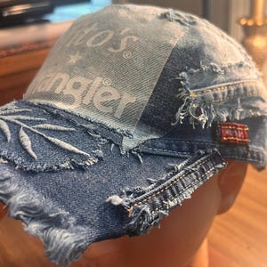May include: Distressed denim baseball cap with frayed edges. Features a patch with the Wrangler logo and a small red pin. The brim has embroidered leaf details. A unique, upcycled accessory.