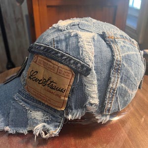 May include: A distressed denim baseball cap with frayed edges and a brown leather patch. The patch features the Levi Strauss signature logo and text. The cap is made of light blue denim and has a curved brim.