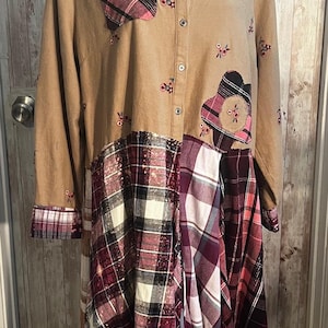 May include: A tan button-down dress with long sleeves and a collared neckline. The dress features plaid patchwork in shades of burgundy, white, and black. Small floral embroidery accents the tan fabric.