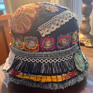 May include: A handcrafted denim bucket hat with a patchwork design. The hat features colorful fabric patches, lace trim, and decorative stitching. The brim is fringed, and the hat has a unique, artistic aesthetic.