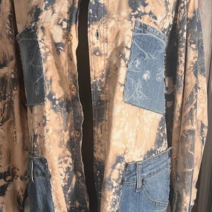 May include: A button-down shirt with a unique design. The shirt features a bleached, tan and navy blue pattern. Denim patches are sewn onto the shirt, including a pocket with a floral design. The shirt has a classic collar and button closure.