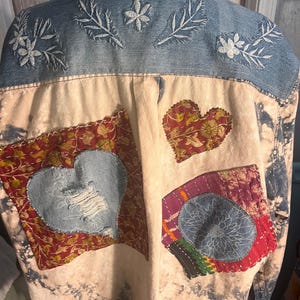 May include: Denim jacket with embroidered floral details on the shoulders. The back features fabric patches with heart and circular designs. The jacket has a bleached, tie-dye effect on the lower portion.