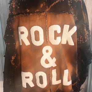 May include: A distressed denim jacket with a brown and black color scheme. The back of the jacket features the words "ROCK & ROLL" in large, white, textured letters. The jacket has frayed edges.
