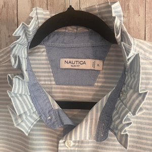 May include: A Nautica slim fit shirt in a light blue and white striped pattern. The shirt features a prominent ruffled collar detail and a button-down front. The interior neck label displays the brand name Nautica, the fit style, and size XL.