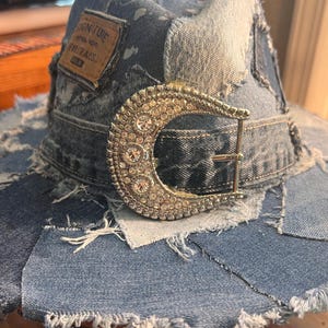 May include: A patchwork denim hat featuring frayed edges and various shades of blue fabric. A large, silver-toned, crescent-shaped belt buckle encrusted with clear rhinestones is attached to the front. A brown leather patch reads SIGNATURE.