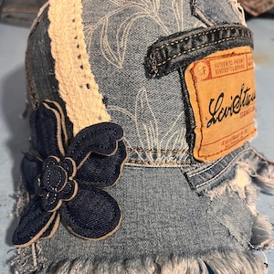 May include: Denim baseball cap with a dark blue flower applique and a cream-colored lace trim. The cap features a distressed denim design with white floral patterns and a brown Levi Strauss label.