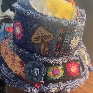 May include: A denim bucket hat with a patchwork design. The hat features a mushroom graphic, crochet flowers, and various fabric patches in shades of blue, yellow, pink, and orange. The brim has a frayed edge.