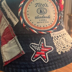 May include: Denim bucket hat with patriotic embellishments. Features a Tito's Handmade Vodka patch, a star-shaped patch with an American flag design, and the text "America the Beautiful."