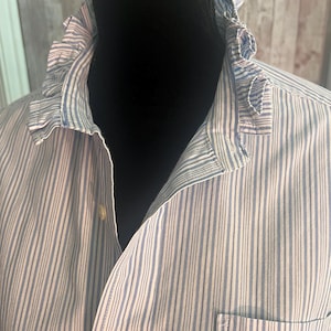 May include: A button-down shirt featuring a light blue and white vertical pinstripe pattern. The shirt includes a distinctive ruffled collar detail, a front button placket with light-colored buttons, and a single chest pocket on the left side.