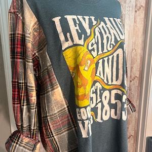 May include: A long-sleeve shirt with a teal body and plaid sleeves featuring a distressed red, black, and white pattern. The front displays a yellow sun graphic with the text LEVI STRAUSS AND CO EST 1853 GOOD VIBES in white, distressed lettering.