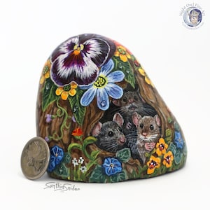 May include: A hand-painted rock depicts three mice in a tree hollow, surrounded by flowers. The rock art features a large purple and white pansy, blue morning glories, and yellow flowers. The artist's signature is visible. The artwork is a unique decorative item.