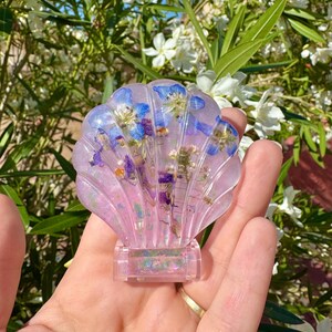 Könnte beinhalten: Ein rosa Muschel-förmiger Papiergewicht aus Harz mit eingebetteten blauen und violetten Blumen und schimmerndem Glitzer.