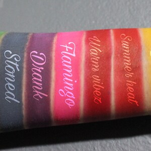 May include: Close-up of a person's arm with swatches of colorful eyeshadow. The swatches are labeled "Cash", "Stoned", "Drank", "Flamingo", "Warm vibes", "Summer heat", and "Sol".